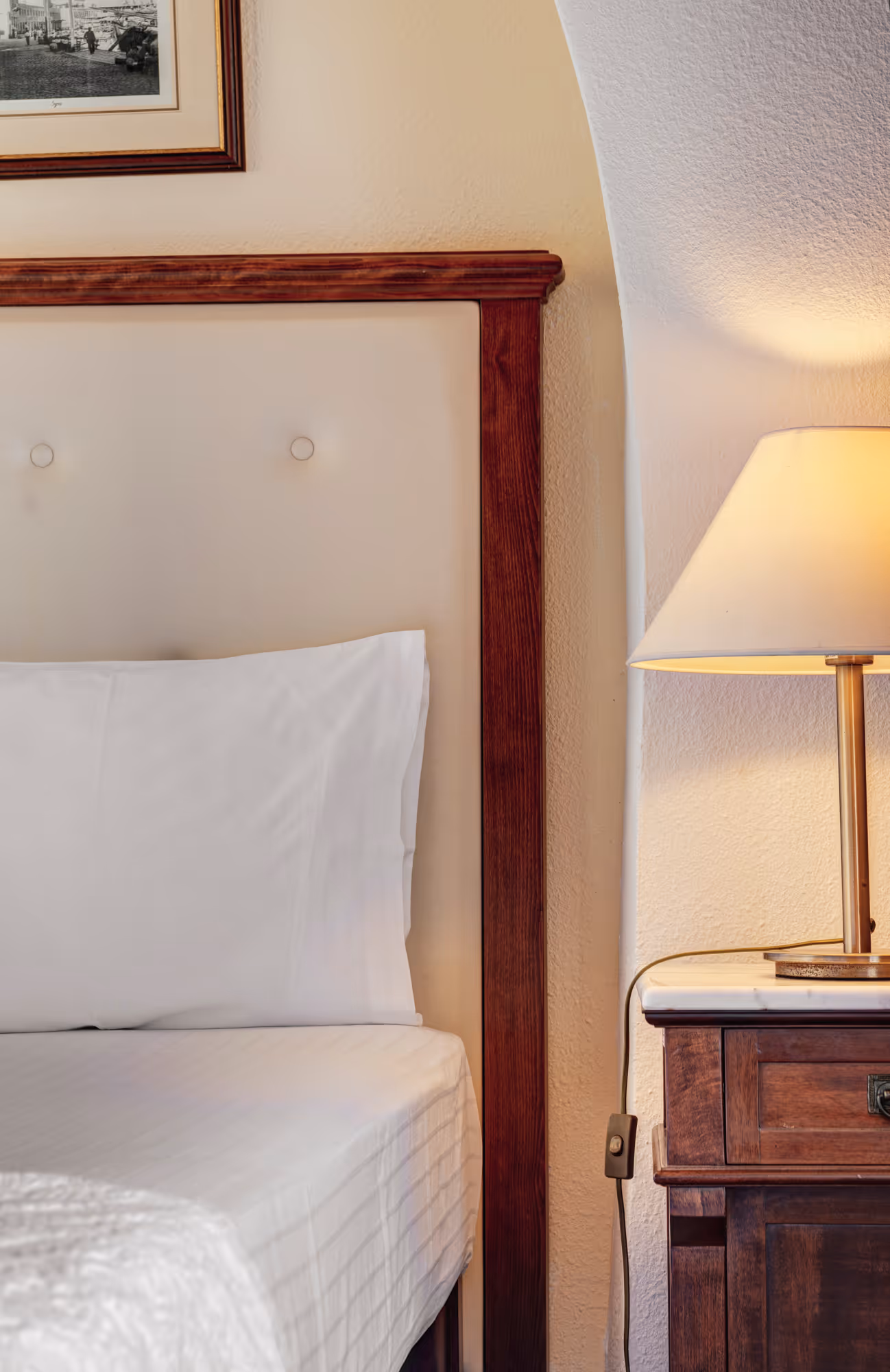 Side view of a bed with white bedding and a wooden headboard next to a wooden nightstand with a lit table lamp.