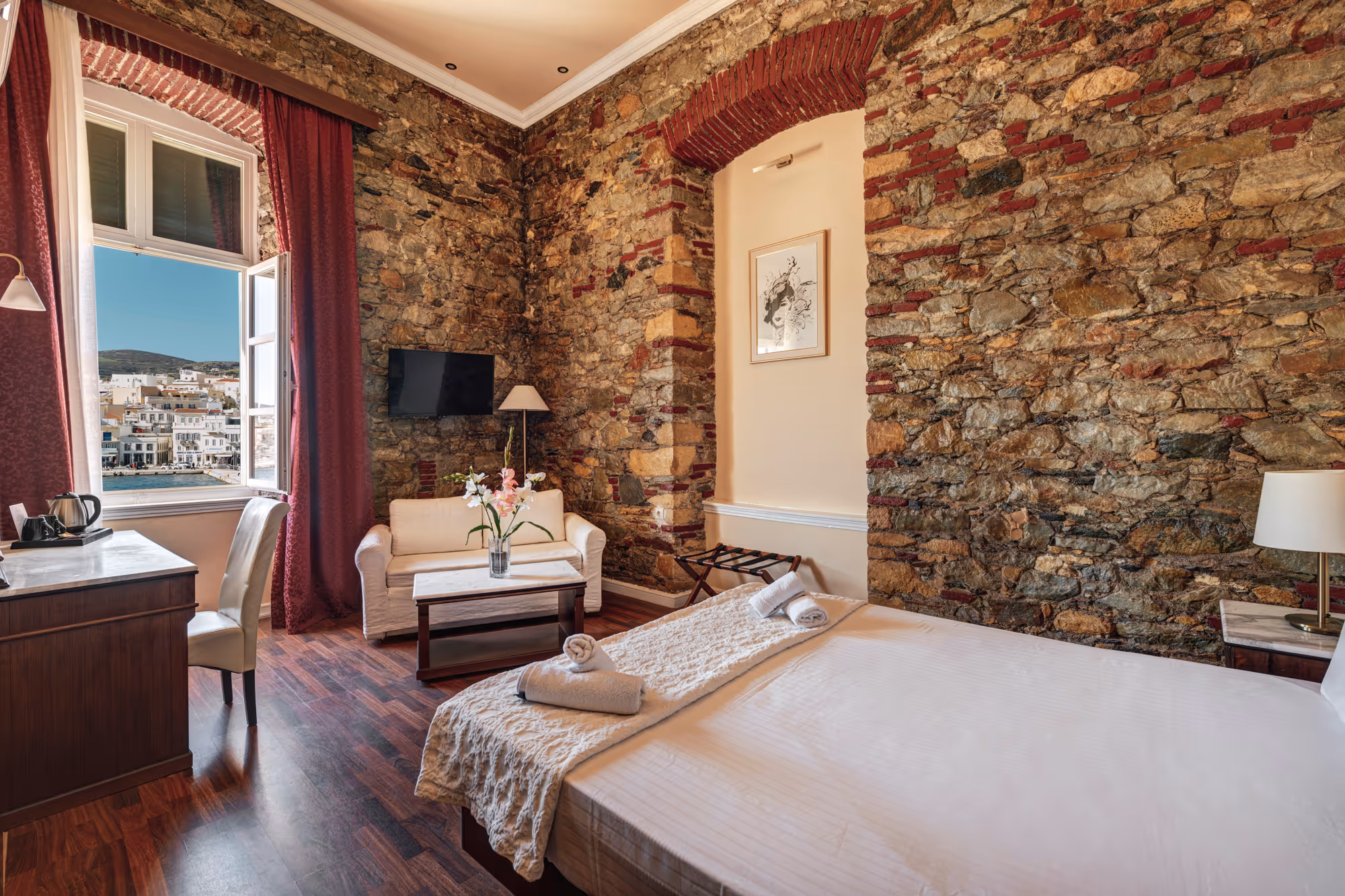 Cozy hotel room with stone walls, a made bed with towels, a white sofa, a small coffee table, desk with chair, and an open window showing a seaside town.