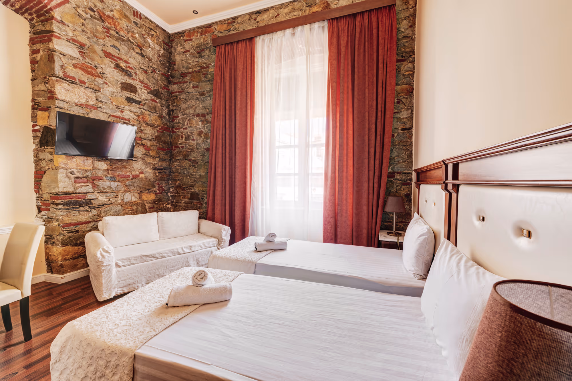 Hotel room with two single beds with white linens, a small white couch, stone accent walls, and a window with red curtains.